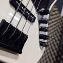 Detailní záběr na spodní část bílé basové kytary s černým pickguardem. Jsou vidět dva černé snímače a černý most s dörti. Na pravé straně jsou umístě…