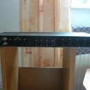 Zadní strana černého rackového zařízení s řadou konektorů. Na panelu jsou nápisy OUT, IN a vpravo MIC/LINE INPUT s šesti kombinovanými konektory. Upr…