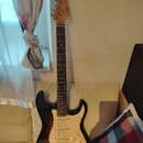 Elektrická kytara s trupem v barevném přechodu od černého k hnědému a krčkem z jasného dřeva s tmavým hmatníkem. Na hlavě je viditelné logo Fender. K…