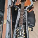 Akustická kytara s hnědým povrchem a cutawayem v černém tvrdém pouzdře s šedým polstrováním. Na hlavě hmatníku je bílý nápis Epiphone a nad ním menší…