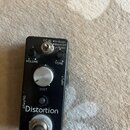Černý efekt pedál s nápisem Distortion. Na horní části jsou ovládací prvky VOLUME, TONE a DIST, přepínač s popisky DC IN, Hi Boost, Boost Off, Lo Boo…