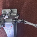 Detailní pohled na kový mechanismus pedálu bicích s řetězem. Části jsou vyrobeny z anodizovaného hliníku v šedé barvě a chromované oceli. Na povrchu…