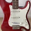 Červená elektrická kytara typu Stratocaster s bílým pickguardem a hnedým hmatníkem. Na spodním snímači je nápis "Dynamic Precision". Kytara má tři bí…