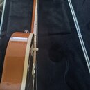 Detailní pohled na hnědou akustickou kytaru v černém polstrovaném kufru. Je vidět bok nástroje s bílým lemováním a vyčnívající bílý knoflík. Na vnitř…