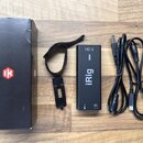 Na dřevěném povrchu leží černá krabice s červeným logem IK Multimedia, černý obdélníkový adaptér iRig HD 2 a černý kabel s konektory. Na adaptéru je…