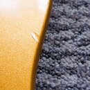 Detailní záběr na hranu nástroje se zlatým metalizovaným lakem. Na povrchu laku je viditelný drobný odřes nebo škrábance. Pozadí tvoří šedý huňatý ko…