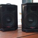 Dva černé reproduktory s kovovými mřížkami umístěné na dřevěném stole. Na spodní části obou reproduktorů je červenobílý nápis APOGEE SOUND INC. Upros…