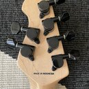 Záběr na hlavu světlé dřevěné kytary s šesti černými ladicími mechanismy. Na zadní straně hlavy je černý nápis MADE IN INDONESIA. Povrch dřeva je lak…