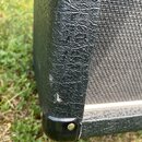 Detailní pohled na roh kytarového komba s černým texturovaným povrchem a šedou tkanou přední mřížkou. V dolním rohu je umístěn černý plastový ochrann…