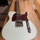 Bílá elektrická kytara s tmelelovým hnedým pickguardem a světlou hmatníkovou deskou. Na kovovém krytu snímače je nápis "TELECASTER" a pod ním menší t…