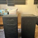 Dva černé, hranaté reproduktory v textilním potahu. Povrch levého reproduktoru vykazuje výrazné pomačkání a deformace materiálu v horní polovině. Na…