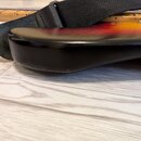 Detailní pohled na spodní hranu korpusu elektrické kytary s barevným přechodem od žluté přes červenou k černé. Na korpusu je nasazení černého textiln…