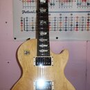 Elektrická kytara s těllem z přírodního světlého dřeva a hnedým hmatníkem. Na hlavě je nápis Gibson. Nástroj má dva humbuckery, čtyři zlaté oválné kn…