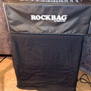 Černý textilní krycí povlak s bílým nápisem ROCKBAG by Warwick. Na horní části povlaku leží kovový obdélníkový box s náborem šroubů a smotaný světle…