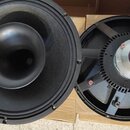Dvě černé reproduktorové jednotky. Levý reproduktor má černou membránu a centrální kompresní horn. Pravý reproduktor je zobrazen zezadu, vidět je kov…