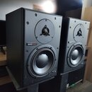 Dva černé regálové reproduktory umístěné na stojanech. Na přední straně každého reproduktoru je nápis dynaudio acoustics v červeném a bílém rámečku.…