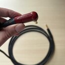 Černý kabel s červeným konektorem, který drží ruka. Na červené části konektoru je vyražen nápis NEUTRIK. Druhý konektor je zlatého odstínu a kabel je…