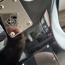 Detail zadní strany černé elektrické kytary s lesklým povrchem, který vykazuje četné škrábance a známky opotřebení. Na těle je přišroubovaná kovová k…