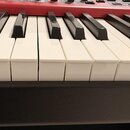 Detailní pohled na klávesnici s bílými a černými klávesami. Horní část instrumentu je červená s několika ovládacími tlačítky. Klávesy vykazují mírné…