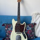 Elektrická kytara s krémovým trupem a hnědým hmatníkem. Na hlavě je nápis "by Taus". Kytara má tmavě červený pickguard s texturou želvy, tři snímače…