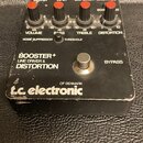 Černý efekt pedál s nápisy BOOSTER+ LINE DRIVER & DISTORTION a logem t.c. electronic OF DENMARK. Horní panel obsahuje čtyři červené oválné knoflíky s…