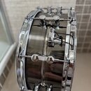 Detailní pohled na stranový rám chromovaného bubnu s tmavým, pravděpodobně akrylovým korpusem. Na kovovém prvku v horní části je vidět nápis "TAMA".…