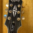 Černý hlavník elektrické kytary s bílým lemováním a zlatými ladičkami. V horní části je bílý nápis Epiphone a uprostřed bílý kosočtvercový ornament.…