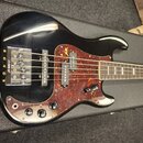 Černá elektrická basová kytara s pěti strunami a hnědým hmatníkem. Nástroj má červeně vzorovaný pickguard a tři černé snímače, z nichž na spodním je…