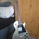 Stříbrná elektrická kytara s černým pickguardem a dvěma humbuckery. Na hlavě hmatníku je viditelné logo Gibson. Nástroj stojí na černém stojanu v int…
