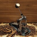 Fotografie zobrazuje kovový pedál k bicím v šedé barvě. Pedál má mechanickou konstrukci s kovovou základnou a horním beaterem s bílým plstěným hřávem…