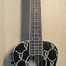 Černá ukulele s bílým vzorem stylizovaných lidských postav na korpusu. Na hlavě nástroje je bílý nápis Fender. Nástroj má čtyři struny a hnědý hmatní…