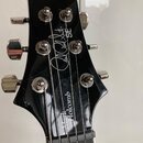 Černá hlava elektrické kytary s chromovanými ladičkami. Na povrchu je bílý nápis „Paul Reed Smith SE“ a vertikální nápis „Holcomb“. Lak vykazuje drob…