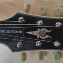 Detail hlavy elektrické kytary v černé barvě s bílým lemováním. Na hlavě je nápis JT-50CUSTOM, logo ve tvaru stylizovaného rohu a svislý nápis Jay Tu…