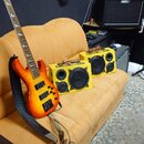Elektrická basová kytara s tělem v oranžovo-žlutém sunburst provedení a hnedým hmatníkem. Vedle ní stojí dva žluté kompaktní reproduktory s nápisem C…