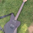 Černá elektrická kytara s matným povrchem a dřevěnou hlavou, položená na trávě. Na snímačích je viditelný nápis "MUSIC". Kytara má dva humbuckery, je…