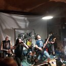Fotografie zachycuje kapelu během koncertu v prostorách s klenutým stropem. V pozadí visí bílý banner s nápisem THE BLACK HEART. Jsou vidět dva kytar…