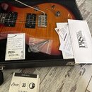 Elektrická kytara s tmeňem v barvě sunburst uložená v černém tvrdém kufru. Na kytaru jsou vidět dva humbuckery, stříbrný most a páka vibrata. V kufru…