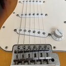 Detail elektrické kytary s bílým pickguardem a tělem v barevnosti sunburst. Na bílém oválkovém knofíku je nápis VOLUME a stupnice 1–10. Kovový kobyln…