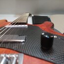 Detailní pohled na tělo a hmatník elektrické kytary s červenohnědým dřevěným povrchem. Nástroj má černý plech s texturou karbonu, bílý snímač u krku…