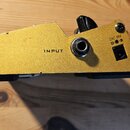 Detail zadní strany žlutého efekt pedálu z kovu. Na šasi je vidět nápis INPUT u vstupního jacku a DC 9V s ikonou polarity u napájecího konektoru. Pov…