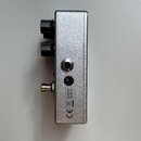 Stříbrný efektový pedál z kovu s dvěma černými ovládacími knoflíky. Na boku jsou umístěny dva konektory, z nichž jeden je napájecí s nápisem 9V a ved…