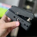 Černý, kompaktní kousek elektroniky v texturovaném plastovém nebo lakovaném šasi s víkem. Na boku jsou vidět dva malé černé oválné přepínače a jeden…
