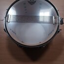 Fotografie znázorňuje bílý buben s kovovým obručem a napnutou blanou. Na blaně je černý nápis REMO WEATHERKING AMBASSADOR SNARE. Přes blanu je nataže…