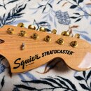Detail hlavy elektrické kytary z přírodního světlého dřeva. Na dřevě je černý nápis Squier BY FENDER STRATOCASTER®. Kytara má zlaté ladičky a stálý m…