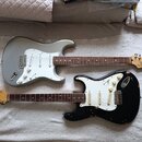 Dvě elektrické kytary s tvarem Stratocaster položené vedle sebe na světlé podložce. Horní kytara je stříbrná s bílým pickguardem a hnědým hmatníkem,…