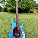 Modrá elektrická basová kytara s čtyřstrunným hmatníkem z tmavého dřeva a naturální hlavou. Tělo má v oblasti snímačů tmavý červený pickguard s textu…