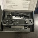 Černý audio rozhraní s nápisem MOTU v originální krabici s pěnovou výplní. Na předním panelu jsou dva kombinované konektory s popisky IN 1L a IN 2R,…