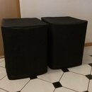 Dva černé, kostkovité reproduktory nebo subwoofery potažené látkou. Jsou umístěny vedle sebe na bílo-černém dlažbovém povrchu. Na přístroji nejsou vi…