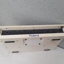 Bílé elektronické zařízení značky Roland s ovládacím panelem na zadní straně. Panel obsahuje několik konektorů, ovratovací knoflík VOLUME a nápisy: P…
