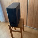 Černý hranatý subwoofer umístěný na dřevěném stoličce. Na spodní části přední strany je kulatý otvor a pod ním bílý nápis LG. Povrch přístroje je hla…